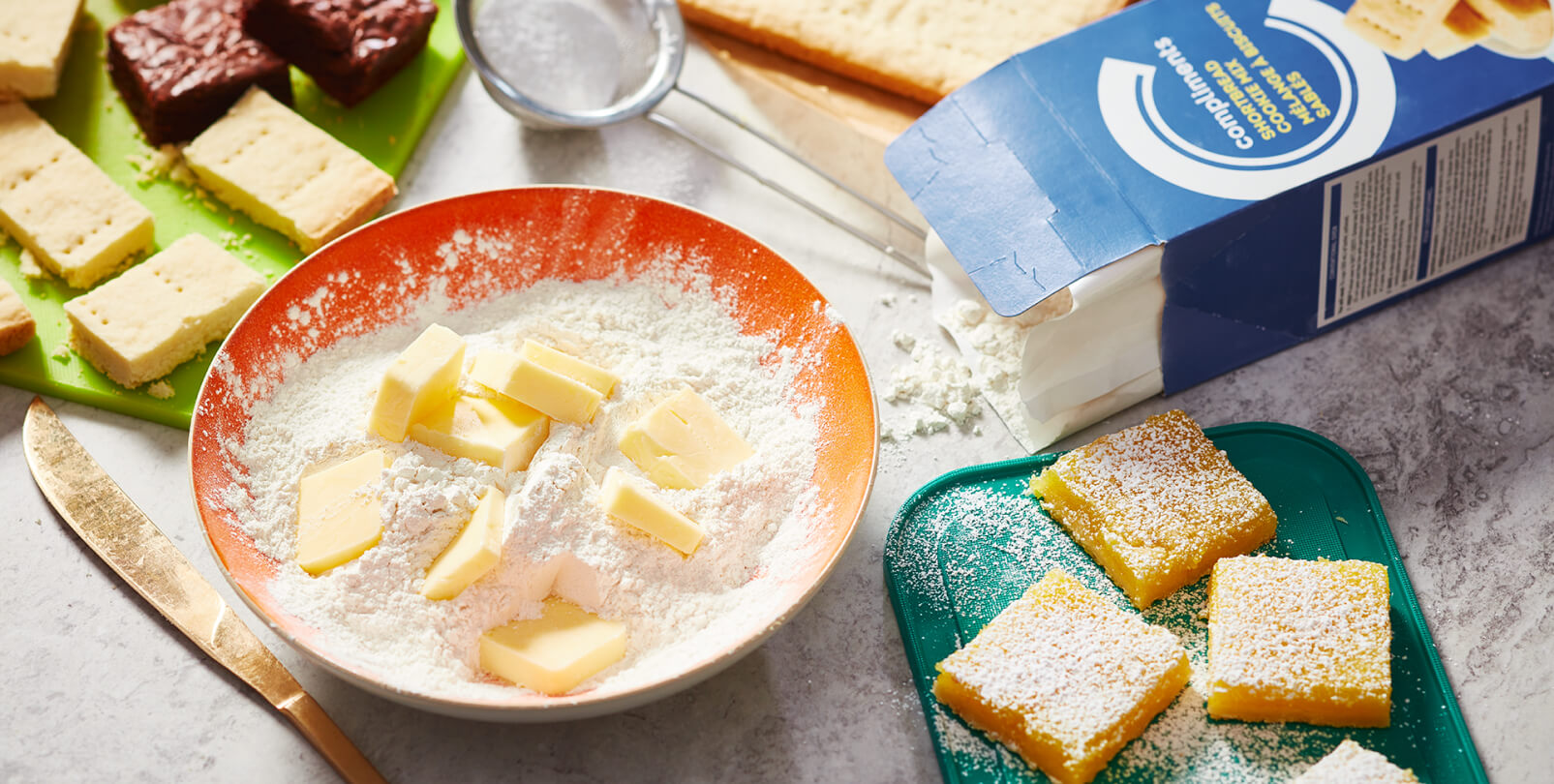 Marble surface with box of Compliments Shortbread cookie mix, baked lemon squares and a mixing bowl of cookie mix and butter.