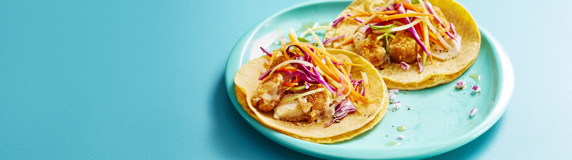 Fond turquoise et assiette blanche avec deux tacos de poisson garnis de salade de chou et carottes.