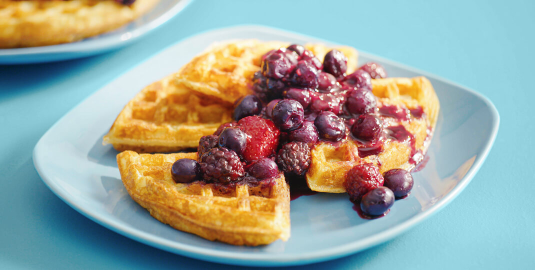 Plateau de table bleu avec une assiette de petit-déjeuner bleue contenant une gaufre cuite nappée d'une sauce aux fruits mixtes.