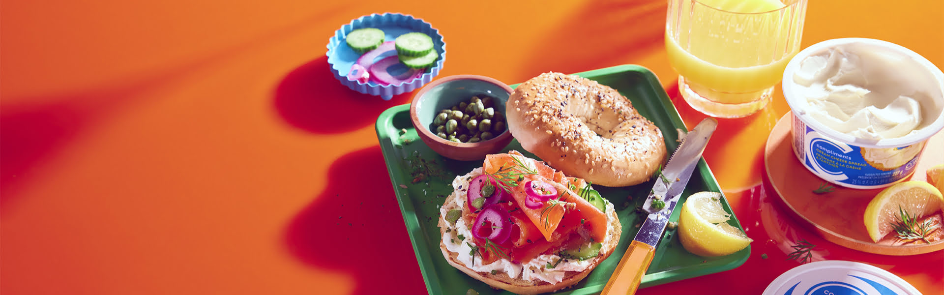 Orange background with breakfast setting including juice, tea, a cream cheese-spread bagel with smoked salmon, dill and capers.