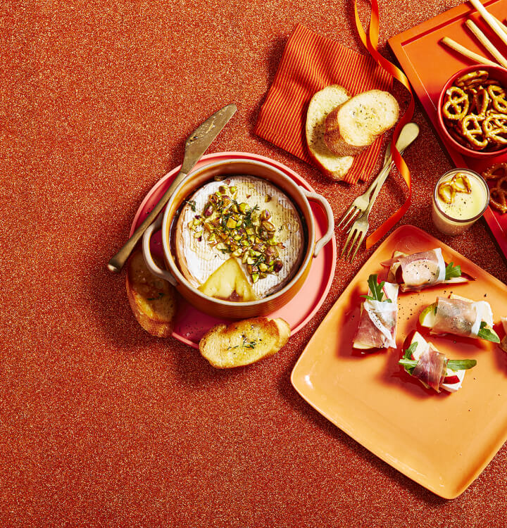 Brie cuit au four garni de noix concassées et d'un accompagnement de crostinis sur une assiette rose, et bouchées de brie et de poire sur un plateau de service orange.