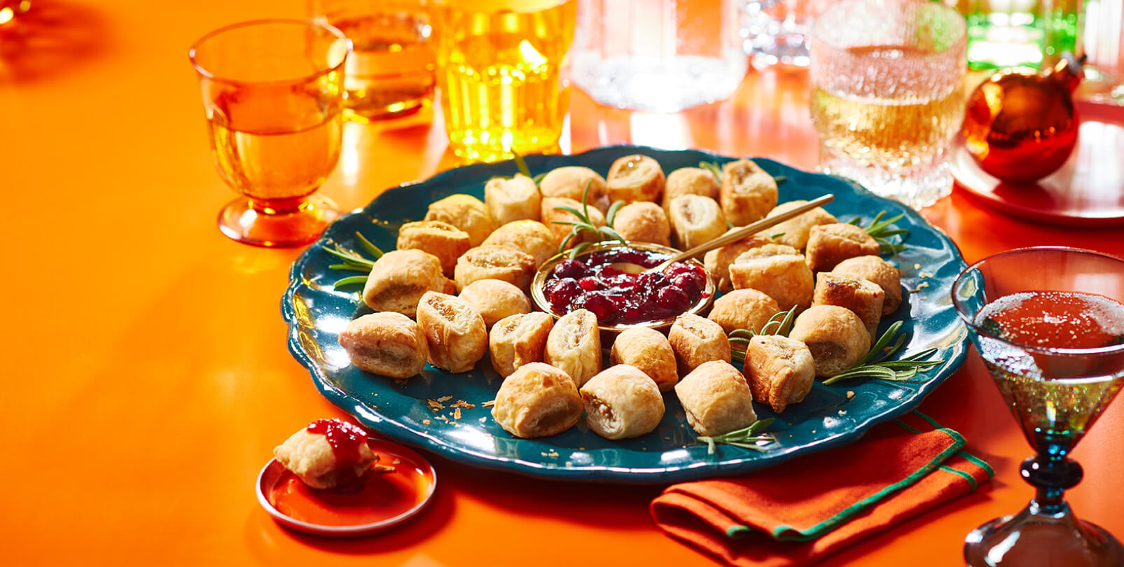 A blue plate of sausage rolls arranged in a wreath shape with fresh rosemary and dipping sauce.