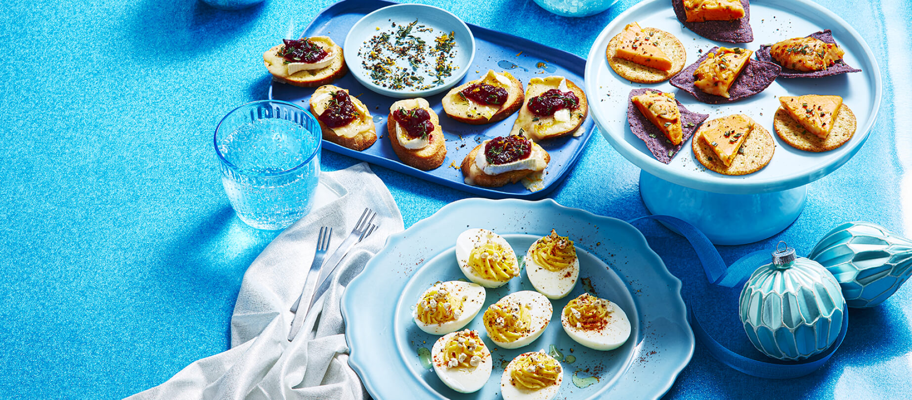 Cheese ball 'n' chips on a white cake stand, brie 'n' cranberries on a blue serving tray and devilled eggs 'n' cheese on a blue plate.