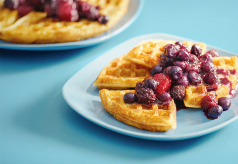 Blue tabletop with a blue breakfast plate with a cooked waffle topped in mixed berry fruit sauce.