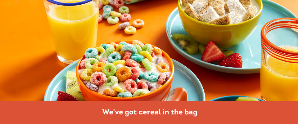 Open bag of Fruity Hoops cereal in background with glass of orange juice and Fruity Hoops cereal in a bowl.